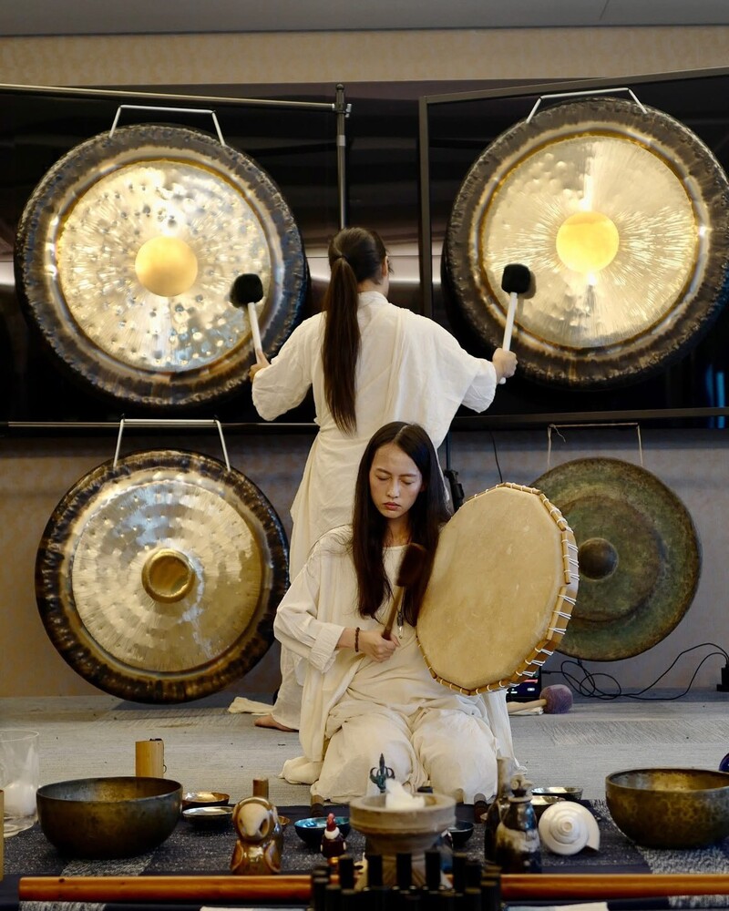 兩位身穿白色服裝的表演者在進行聲音療癒儀典，一位站立敲擊大型銅鑼，另一位跪坐演奏手鼓，周圍擺放頌缽、樂器與自然元素，呈現沉靜而儀式感十足的場景。
