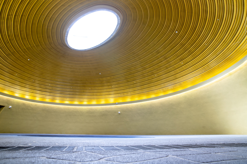 臺灣客家茶文化館的天光大廳室內空間,圓形挑高穹頂中央留有天窗,可透入自然光;金黃色的環形牆面與同心圓天花構成安定而開闊的場域,適合儀典與沉浸式表演。