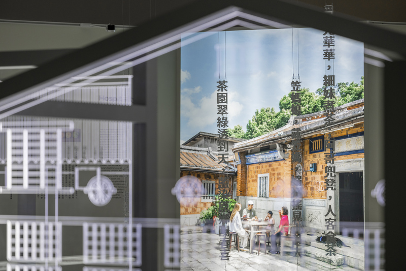 「臺灣客家茶文化館特展《尋客茶路》主視覺，呈現傳統三合院場景，訪客在茶桌前對談飲茶，背景牆面寫有『茶園翠綠透到天』等詩句，展現客家茶生活與詩意文化」