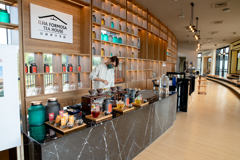 「福爾摩沙茶館內部茶吧台,工作人員現場示範熱茶沖泡與手沖流程,背景為整面茶罐展示牆,環境設計現代且具文化感,地點位於桃園龍潭的臺灣客家茶文化館